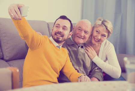 Smiling young son doing selfie with mother and grandfather at home at tableの写真素材