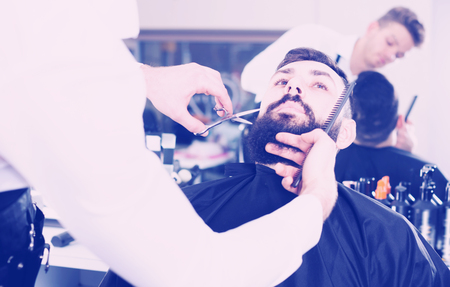 Smiling guy stylist creating new shape for beard of client at hairdressing salonの写真素材