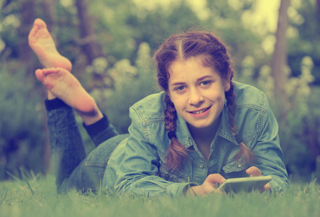positive young girl using digital tablet while lying in green spring  gardenの写真素材