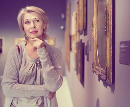Glad cheerful positive mature woman standing in art museum near the painting in baguetteの写真素材