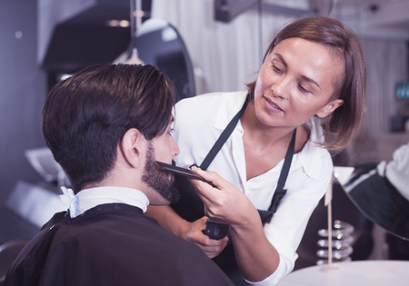 portrait of woman hairdresser shaving man's beard in barbershopの写真素材