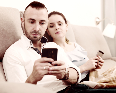 Man ans his wife are chatting on phone in time resting on sofa at home.の写真素材