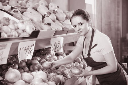 Young positive smiling female seller putting fresh goods on shelves in greengroceryの写真素材