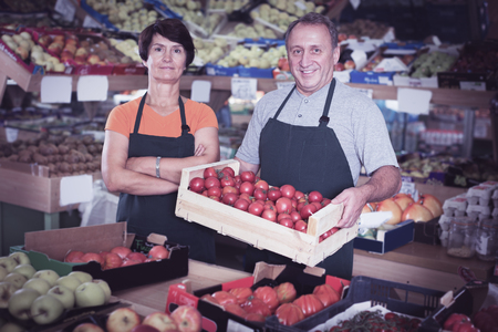Smiling couple sellers is standing with box with tomatos in the food storeの写真素材