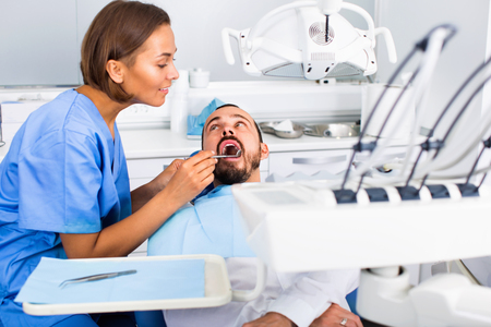 Glad positive stomatologist girl in uniform is taking visional inspection of a man on the chair in dental clinic.の写真素材