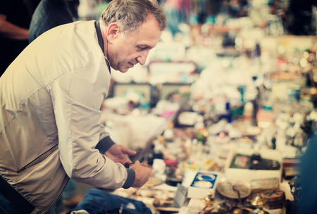 Positive glad cheerful elderly man choosing interesting souvenirs at traditional flea marketの写真素材