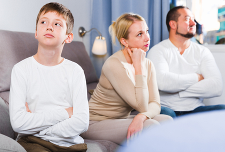 Upset son and frustrated parents sitting on sofa and looking away after quarrelの写真素材