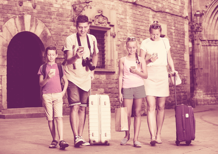 Young family of four with suitcases walking along city using phonesの写真素材