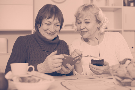 Two positive senior women using phones together over cup of teaの写真素材