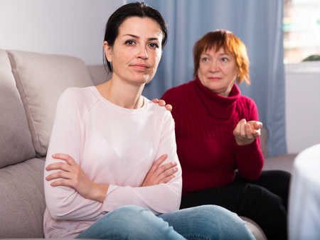 Sad mature female and daughter sitting at sofa after quarrel at homeの写真素材