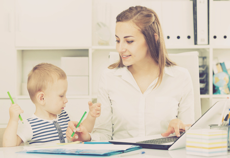 Female employee is working on laptop while child painting on papers in office.の写真素材