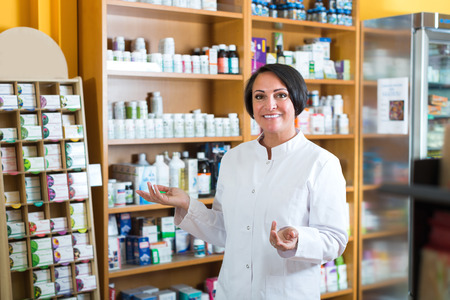Glad mature female seller in uniform standing with dietary supplements in pharmaceutical  storeの写真素材