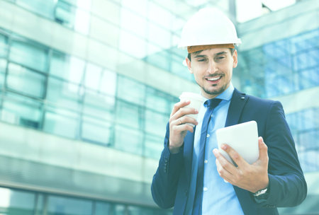 Happy cheerful positive smiling man in helmet with touchpad and coffee in the hand