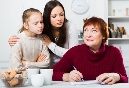 Serious senior woman signing documents while disgruntled daughter and granddaughter staring intently