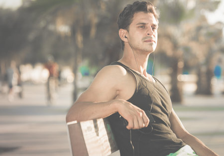 Handsome sporty man resting after workout sitting on bench in earphones