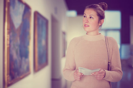 Young woman visitor with guide book looking at exposition in art museum