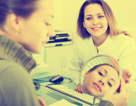 Young positive woman looking at mirror during consultation with doctor in aesthetic medicine centerの写真素材