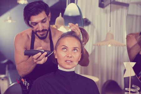 Positive sexy adult hairdresser working with hair clipper with woman の写真素材
