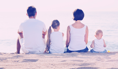 Glad cheerful man and woman with two kids sitting with back to camera on sandy beachの写真素材