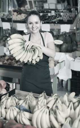 Portrait of young smiling spanish saleswoman with cluster of yellow bananas in storeの写真素材