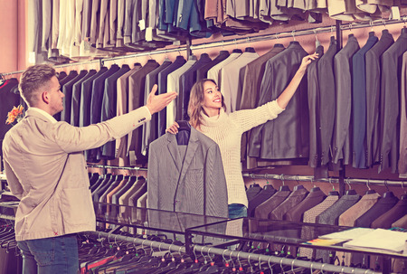 Young cheerful positive woman seller assisting man in choosing suit in mens cloths storeの写真素材