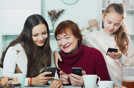 Happy grandmother, mother and daughter using mobile gadgets at table at homeの写真素材