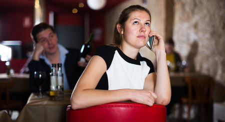 Unhappy woman talking on mobile phone sitting apart in restaurant with drunk man behind の写真素材