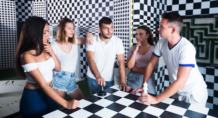 Group of positive adults standing near chessboard in quest room and solving conundrumの写真素材