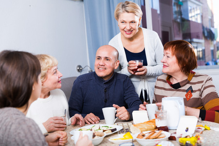 Smiling aged friends are sitting at table with tea and talking about life at home.の写真素材
