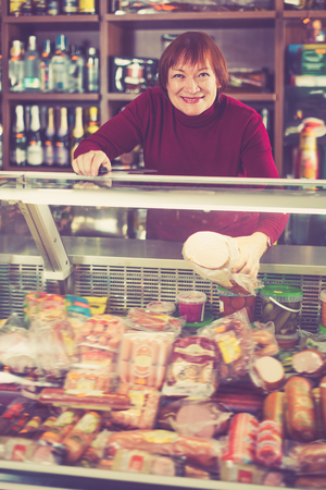 Cheerful female selling sausage and ham at the counter in supermarketの写真素材