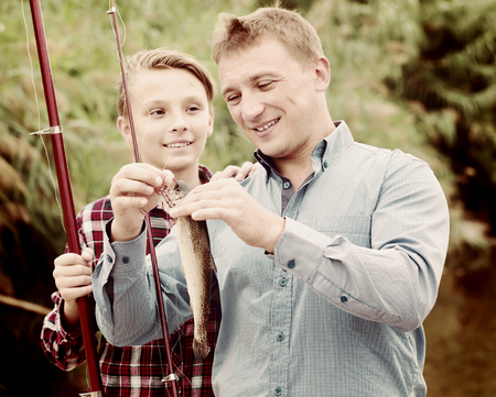 Portrait of laughing man with teenager son looking at fish on hook in hands outdoorsの写真素材