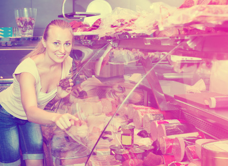 Smiling charming woman customer choosing salami in delicatessen section in food shopの写真素材