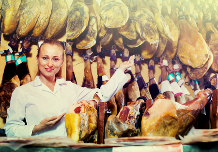 Positive friendly smiling young female seller wearing uniform working with iberian ham in meat shopの写真素材