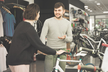 Young cheerful positive man seller demonstrating bicycle to woman client in storeの写真素材