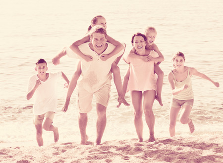 Large friendly  family of six people happily running and carrying kids on parents back on beachの写真素材