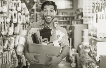 Smiling young man standing near racks in paint store holding basket with toolsの写真素材