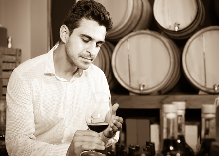 portrait of positive spanish man tasting wine sample in glass in alcohol section with woodsの写真素材