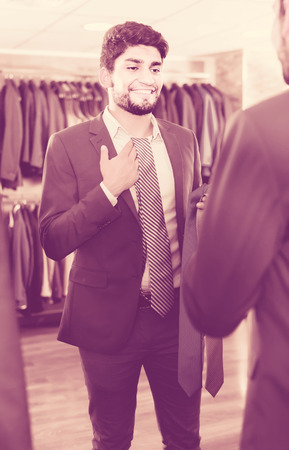 Customer male is picking up tie for jacket in front of the mirror in men's shop.の写真素材