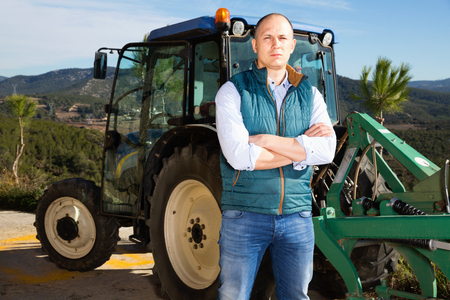 Confident pleasant  male owner of vineyard posing near tractor outdoors in sunny dayの写真素材