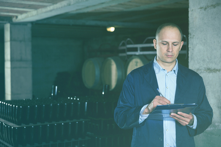 Wine producer with clipboard inspecting wine bottles on racks in wine cellarの写真素材