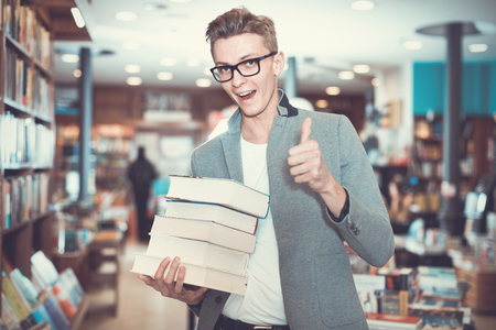 Portrait of happy positive young man holding stack of books in hands in bookshopの写真素材