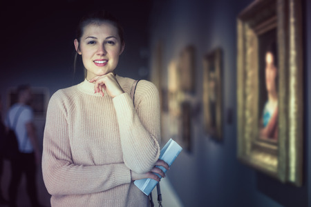 Young female visitor with guide book looking at exposition in art museumの写真素材