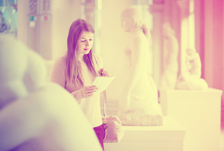 Glad cheerful positive girl looking with interest at ancient sculptures in museum, using guidebookの写真素材
