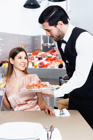 Attractive cheerful  smiling girl served by waiter, enjoying dinner in fish restaurantの写真素材