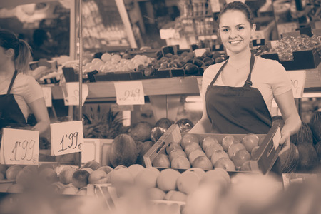 Young happy cheerful positive smiling saleswoman with box of ripe orange persimmons in storeの写真素材
