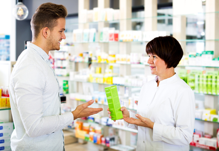 Portrait of glad woman druggist wearing white coat giving advice to customer in pharmacy. Focus on the manの写真素材