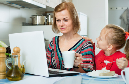 Positive woman sitting with laptop in the kitchen and eating together with her cheerful daughterの写真素材