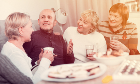 Smiling aged friends are sitting at table with tea and talking about life at home.の写真素材