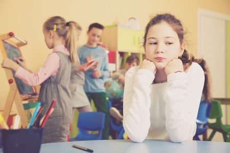 Upset girl sitting at table in schoolroom during break on background with other pupilsの写真素材