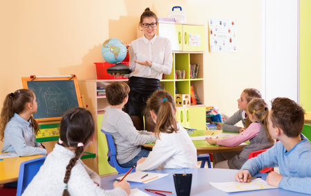 Cheerful female teacher giving geography lesson in classroom, showing pupils globe 